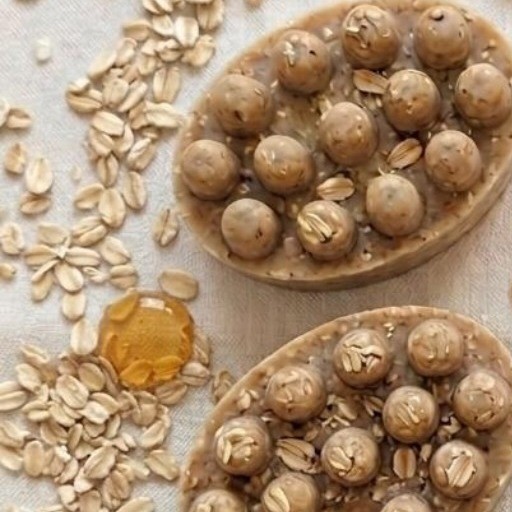 Dos jabones artesanales de avena y miel color tostado, entre copos de avena y gotas de miel sobre lino