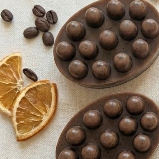 Dos jabones artesanales de café color marrón oscuro, entre granos de café y rodajas de naranja secas