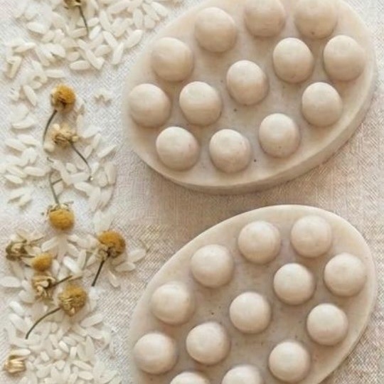 Dos jabones artesanales color crema, rodeados de flores de manzanilla y granos de arroz sobre lino